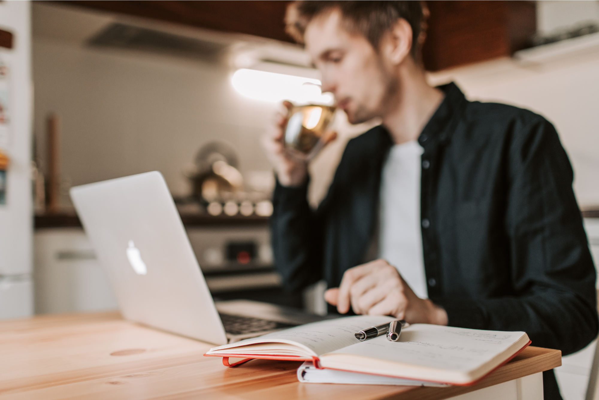 A man sitting at a kitchen table with a laptop, drinking from a mug, and reviewing notes in a notebook — representing lifestyle research on male fertility and environmental sperm health.